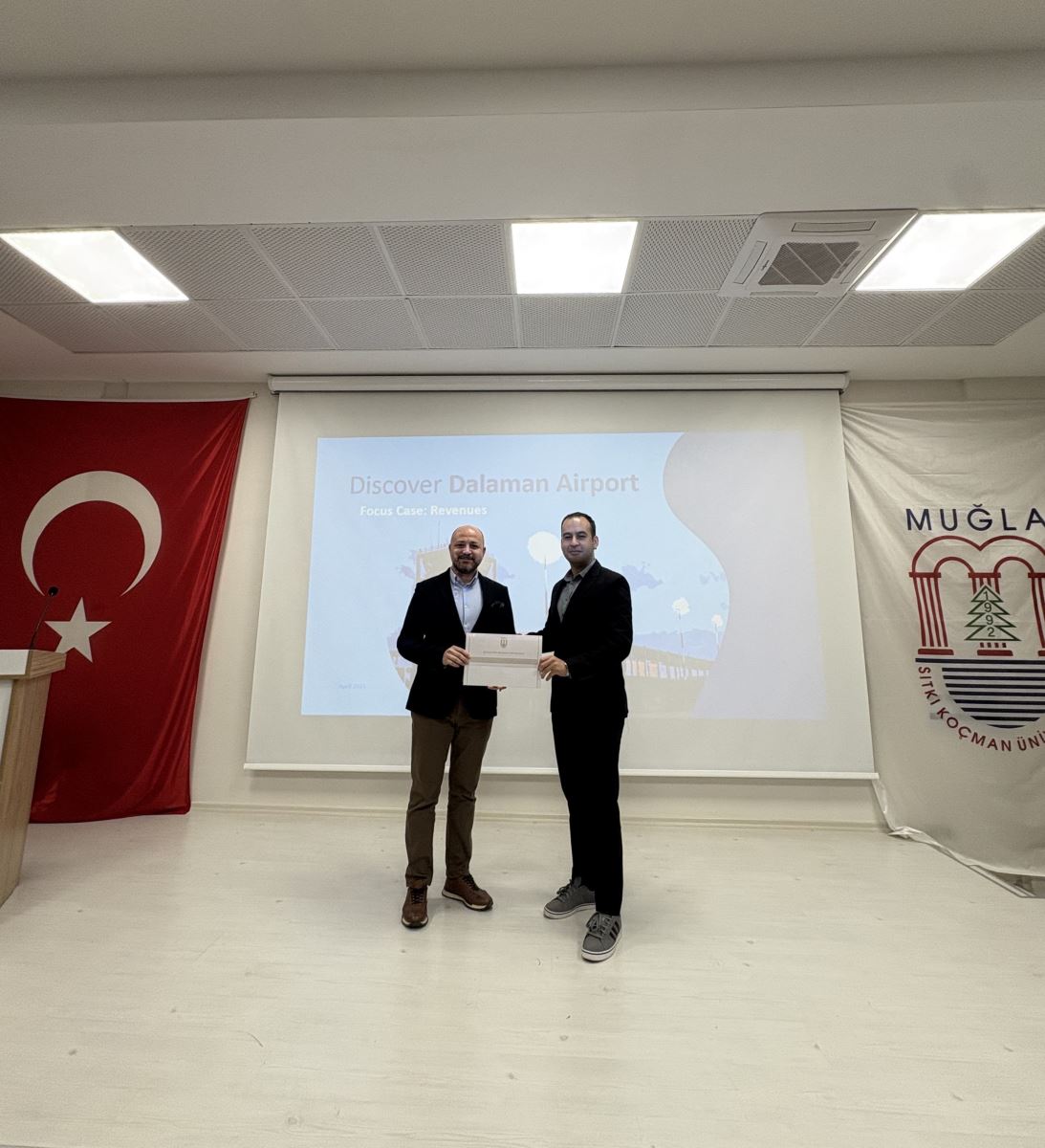 Ahmet Orçun Songur ve bir öğretim üyesi, sahnenin ortasında yan yana duruyorlar. İkisi de gülümseyerek bir teşekkür belgesini birlikte tutuyorlar. Arka plandaki perdede "Discover Dalaman Airport" yazılı bir slayt ve sağ tarafta Muğla Sıtkı Koçman Üniversitesi'nin amblemi yer alıyor.